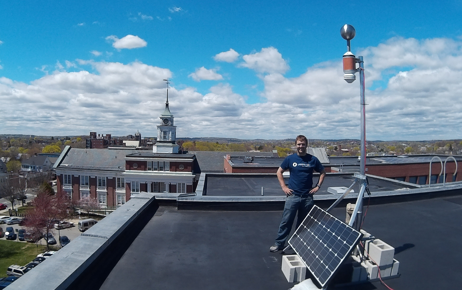 An Understory Weather team member stands in front a solar array hooked to a weather monitoring system on the roof of Somerville High School.